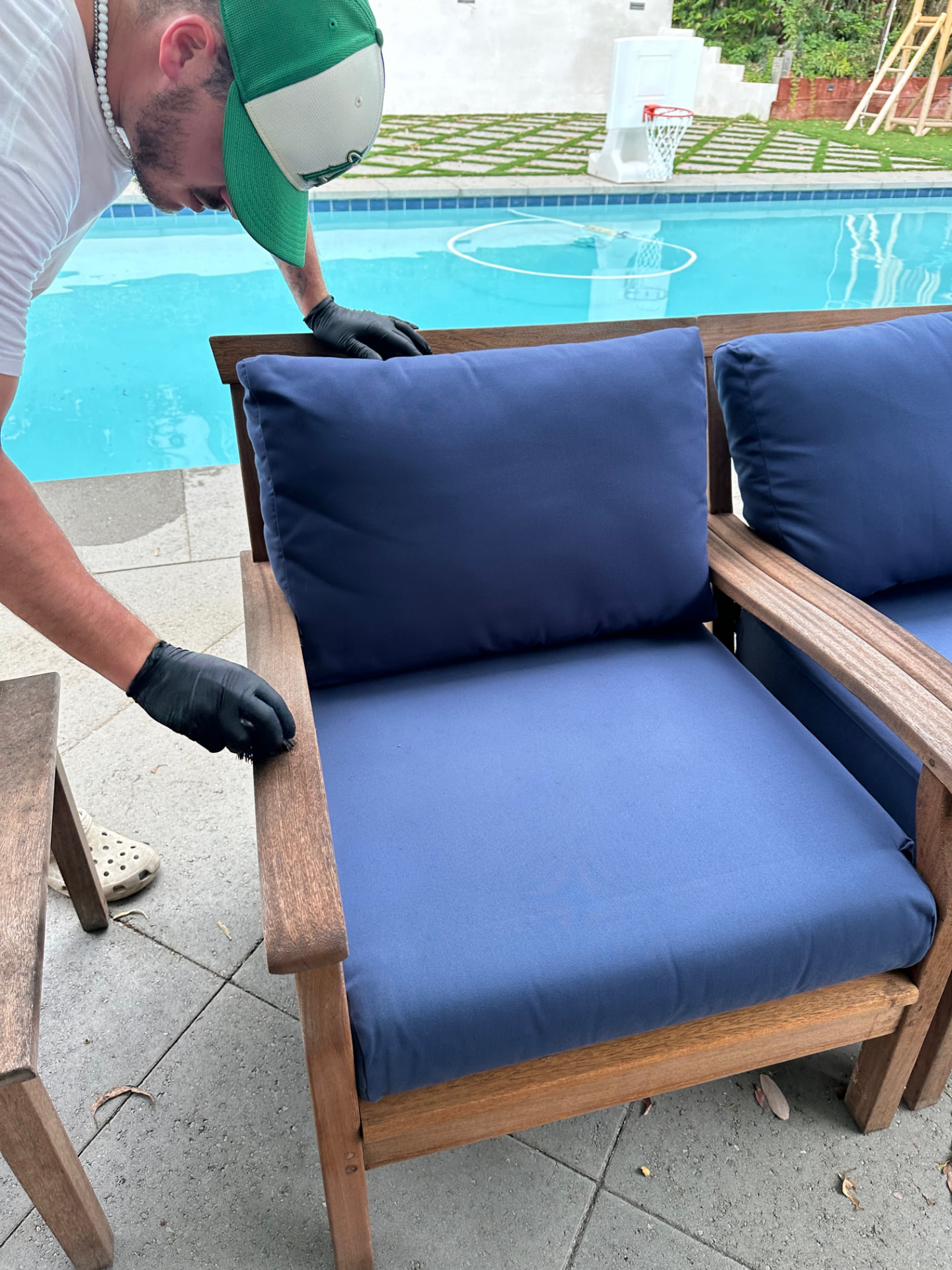 PatioMaid technician cleaning a wooden outdoor sofa with blue cushions by a pool in San Diego